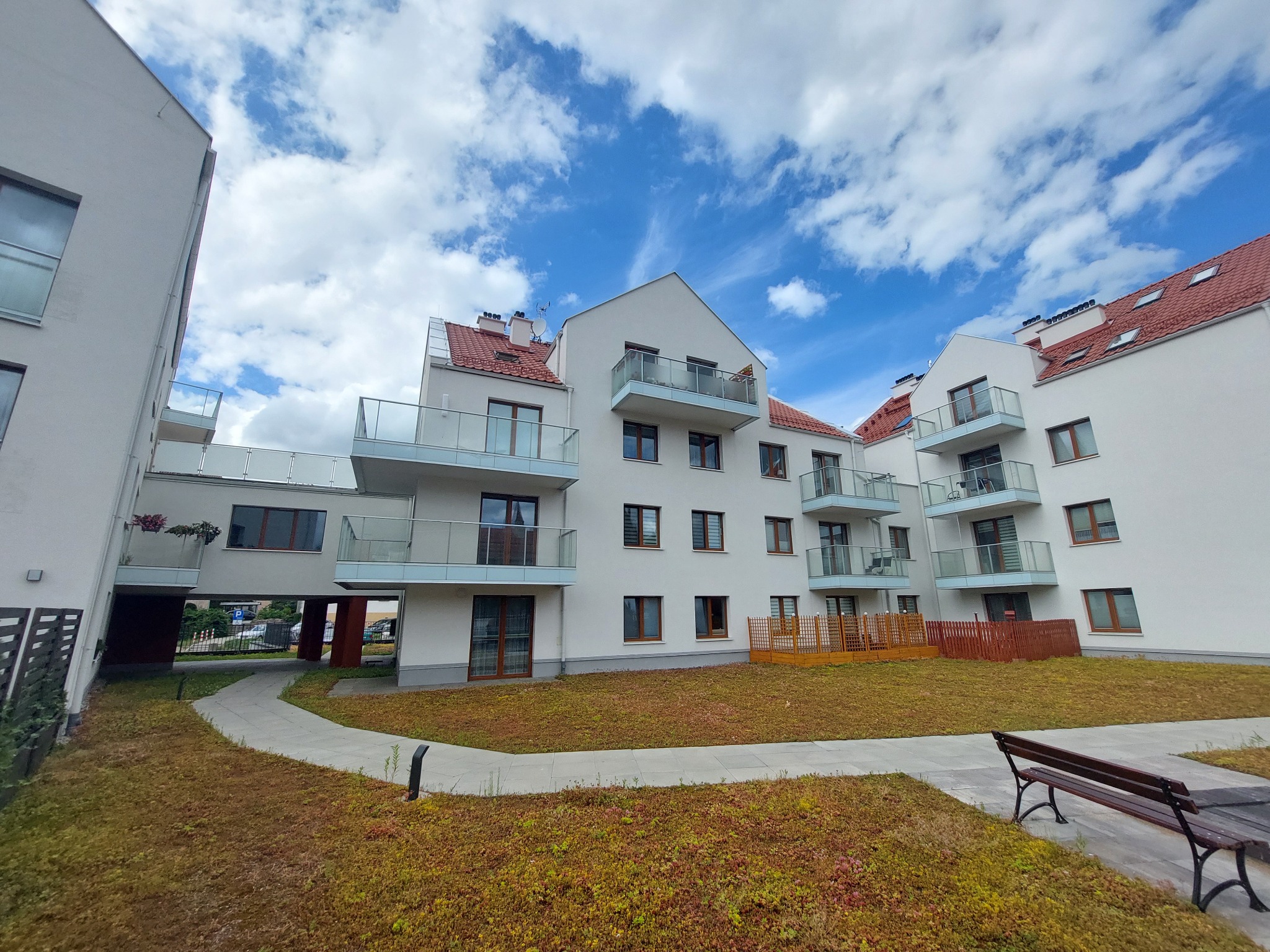 Nowoczesny budynek apartamentowy z balkonami i zielonym dachem na poziomie gruntu, widok z poziomu chodnika, błękitne niebo z chmurami w tle.