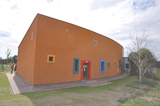 Pomarańczowy budynek o zaokrąglonym kształcie z kolorowymi oknami i czerwonymi drzwiami wejściowymi, widziany z zewnątrz w słoneczny dzień.