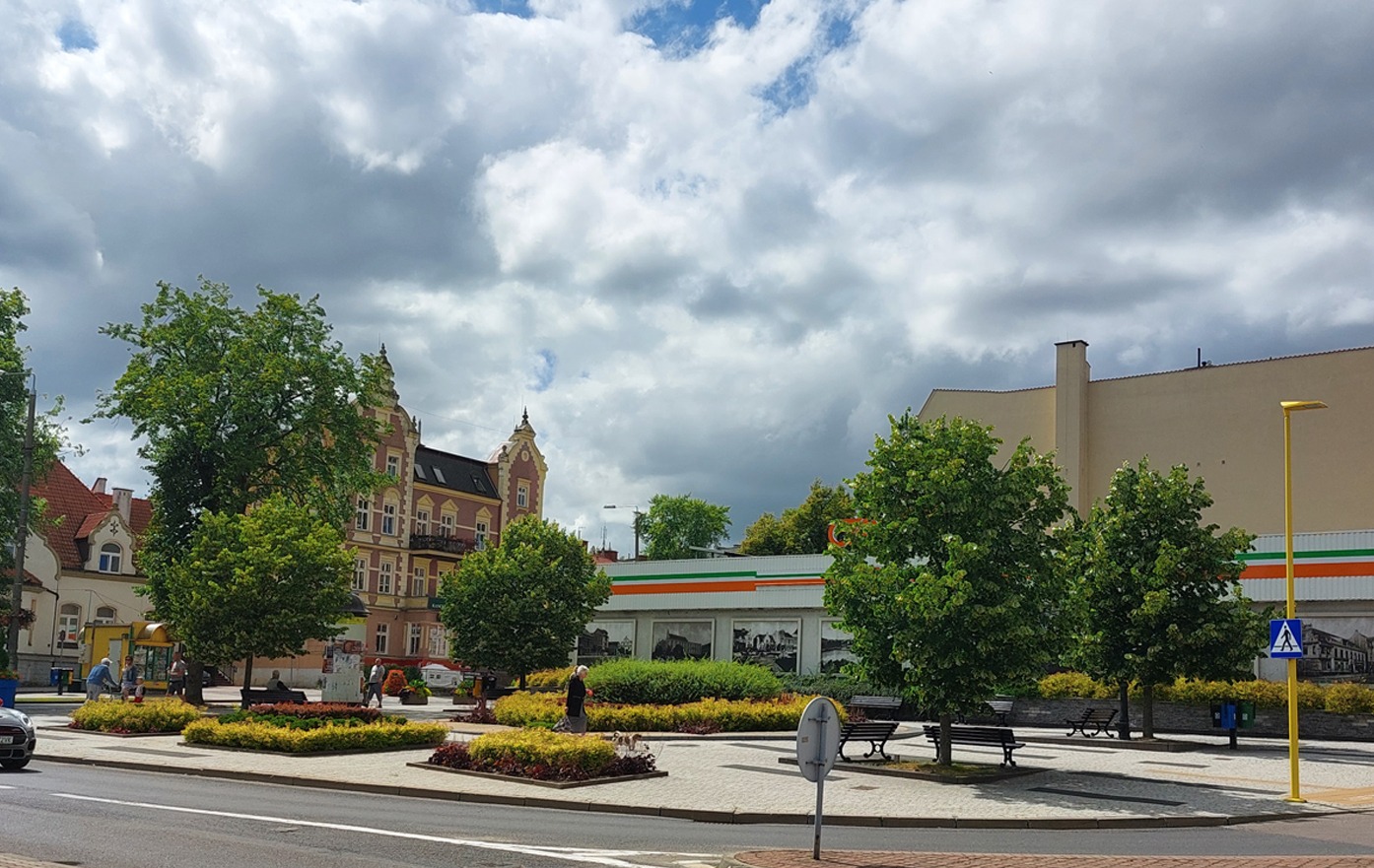Miejski plac z rabatami kwiatowymi, ławkami i drzewami w słoneczny dzień pod pochmurnym niebem.