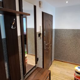 Daniel Grajek Usługi Remontowo-Budowlane - Wnętrze przedpokoju po remoncie: brązowe panele podłogowe ułożone w jodełkę, ściany z szarą strukturą, brązowe meble z lustrem, widoczne puszki z farbą w rogu.