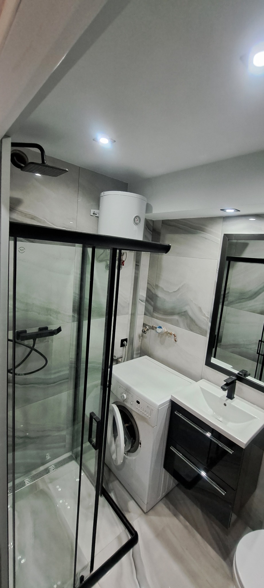 Wykończona łazienka z kabiną prysznicową typu walk-in z czarnymi profilami, pralką, umywalką z szafką w kolorze czarnym i bojlerem na ścianie, płytki imitujące marmur.