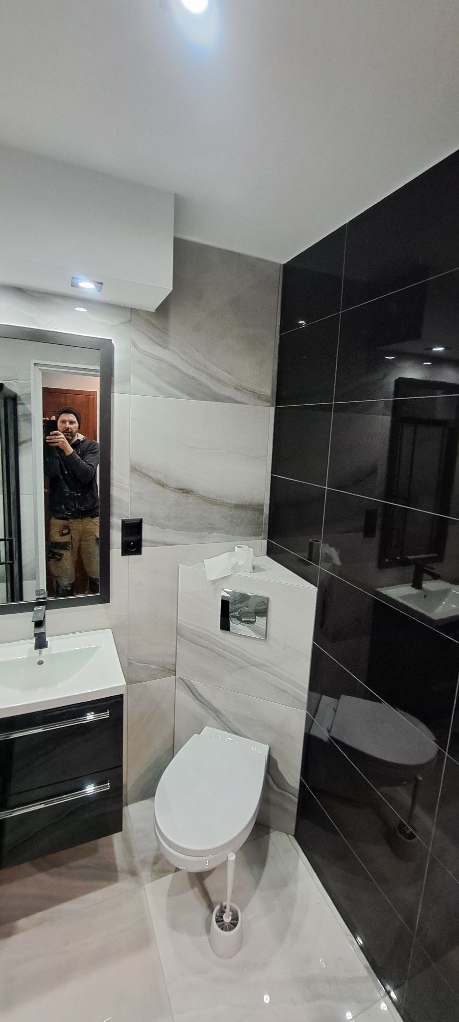 Wykończona łazienka z czarno-białymi płytkami imitującymi marmur, podwieszana toaleta, umywalka z czarną szafką, widoczne odbicie wykonawcy w lustrze.
