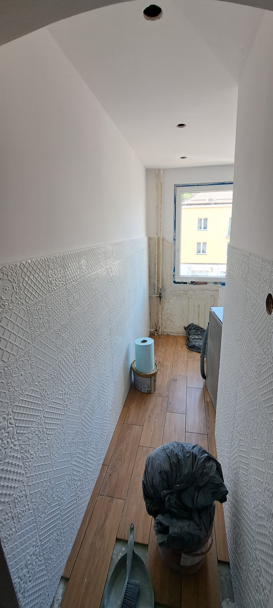 Wąski korytarz w trakcie prac wykończeniowych: położona podłoga z paneli imitujących drewno, ściany częściowo pokryte dekoracyjną białą strukturą, widoczne narzędzia i materiały budowlane, otwory...