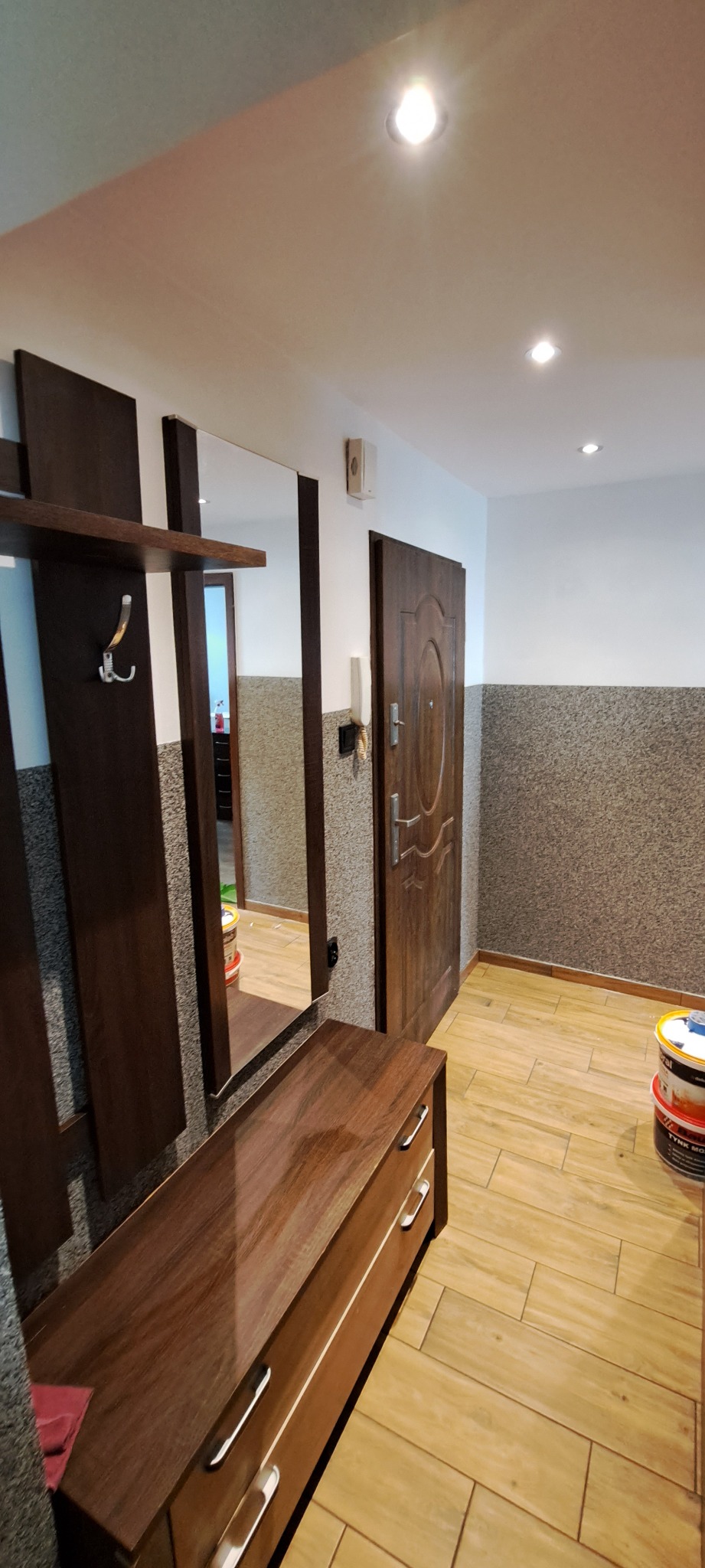 Wnętrze przedpokoju po remoncie: brązowe panele podłogowe ułożone w jodełkę, ściany z szarą strukturą, brązowe meble z lustrem, widoczne puszki z farbą w rogu.