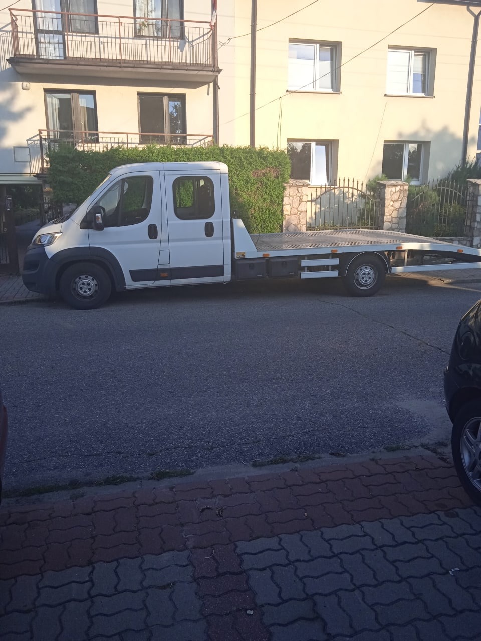 Biały samochód z platformą transportową zaparkowany na ulicy przed budynkiem mieszkalnym z balkonem.