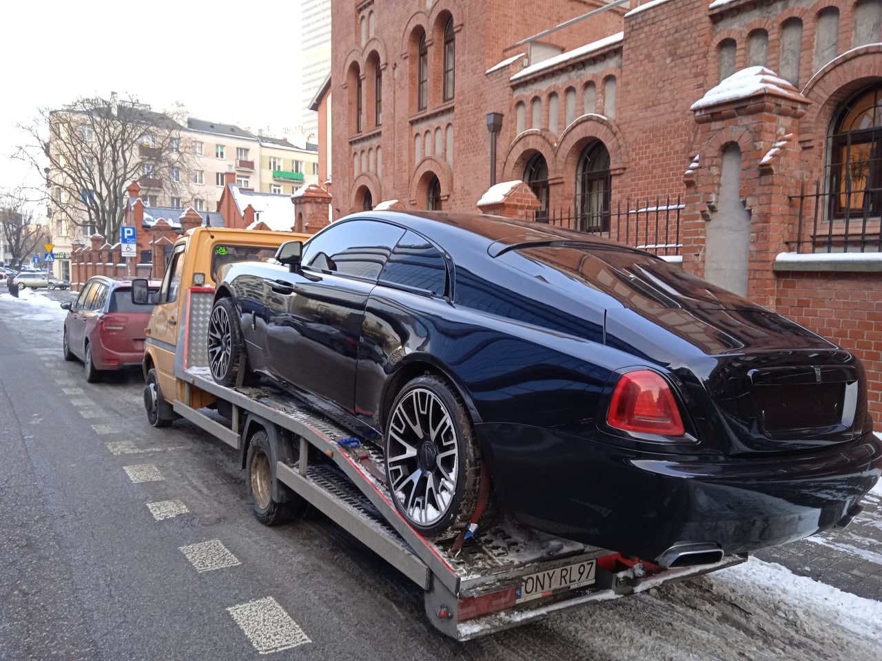 Czarny luksusowy samochód sportowy na platformie pojazdu pomocy drogowej, widok z tyłu, na tle ceglanego budynku z łukowymi oknami i zimowego krajobrazu miejskiego.