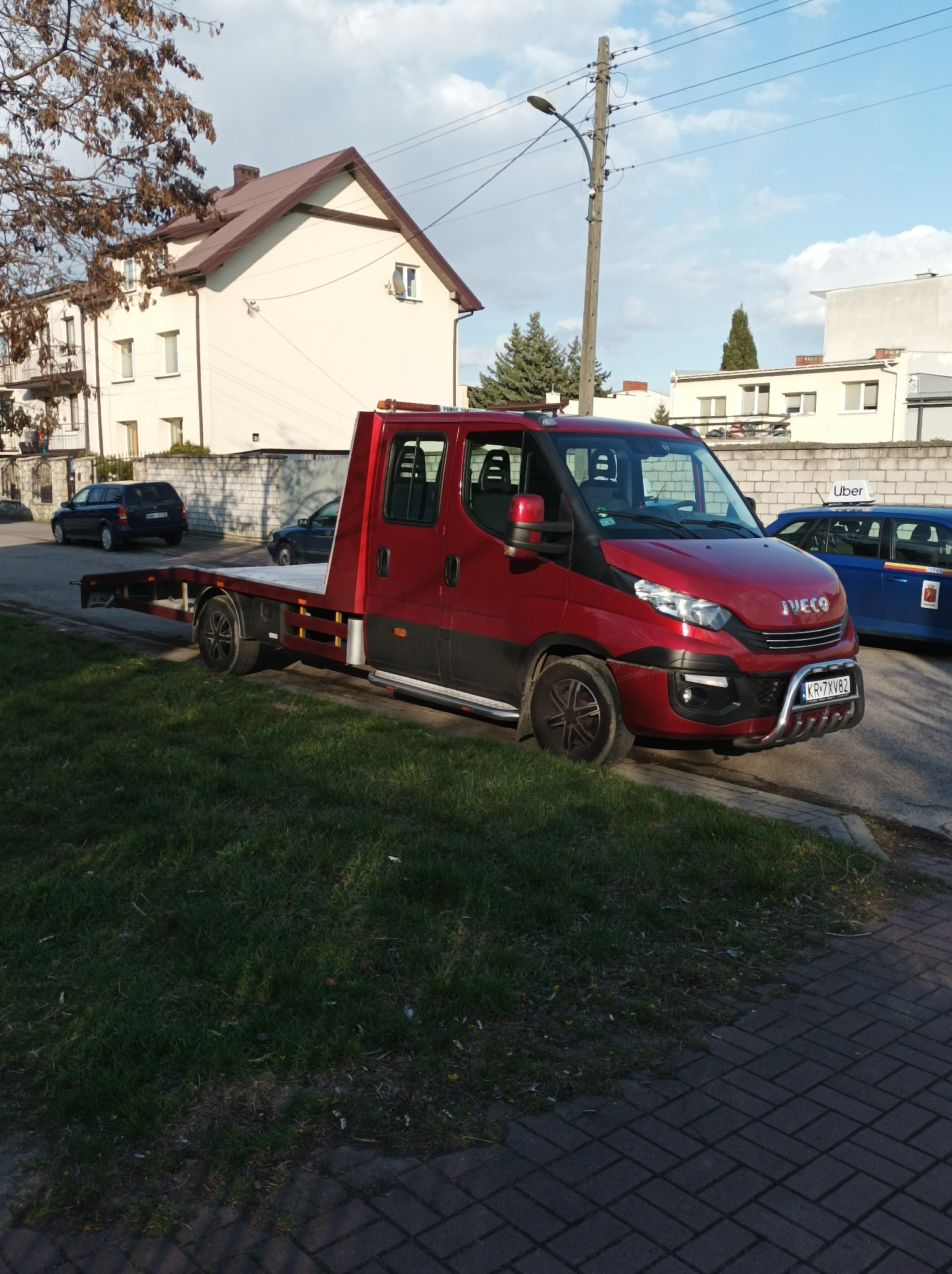 Czerwona laweta Iveco zaparkowana na chodniku obok trawnika, z widocznym numerem rejestracyjnym i budynkami w tle.