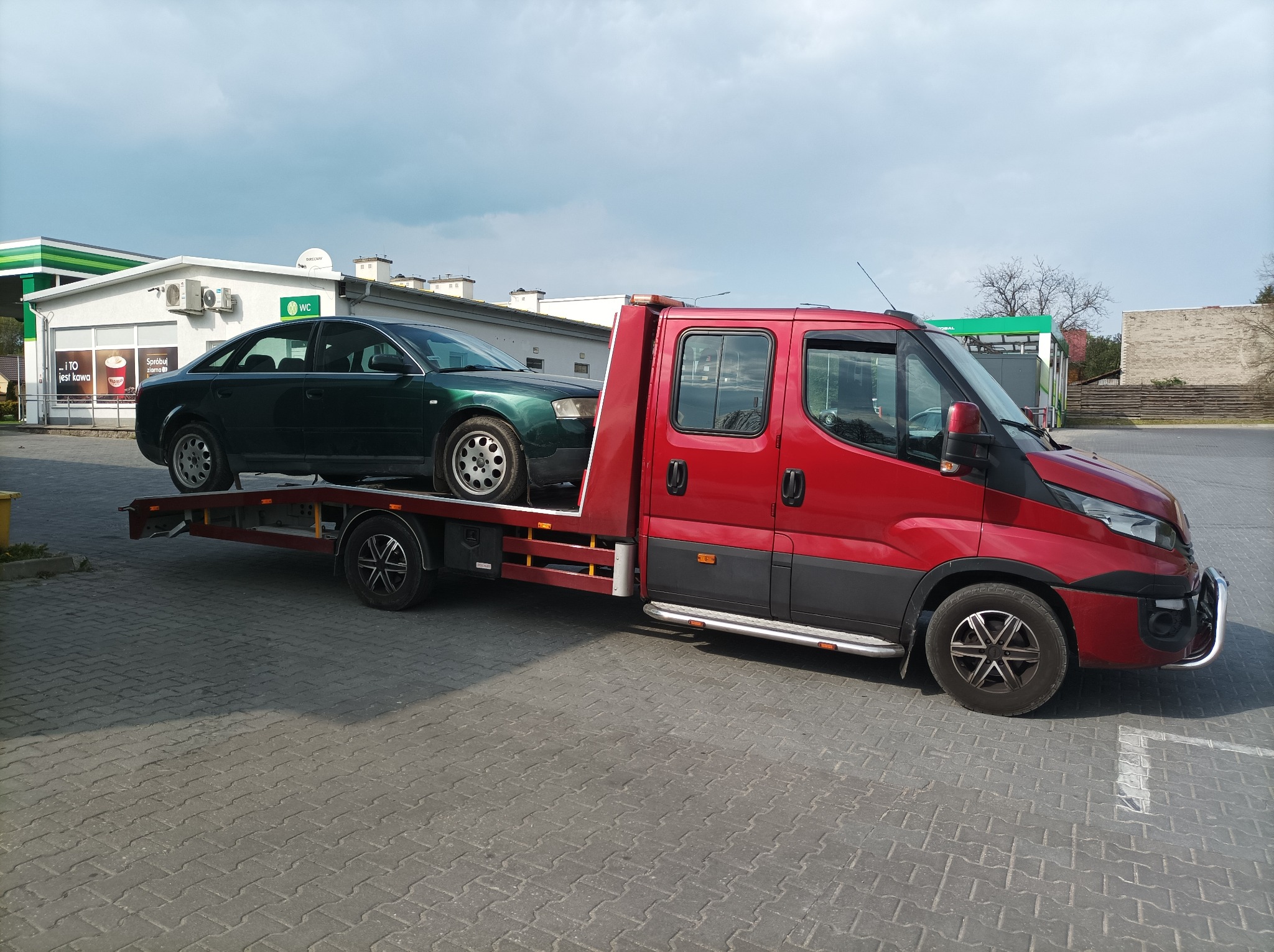 Czerwona ciężarówka z platformą transportową przewozi zielony samochód osobowy na parkingu stacji benzynowej w słoneczny dzień.