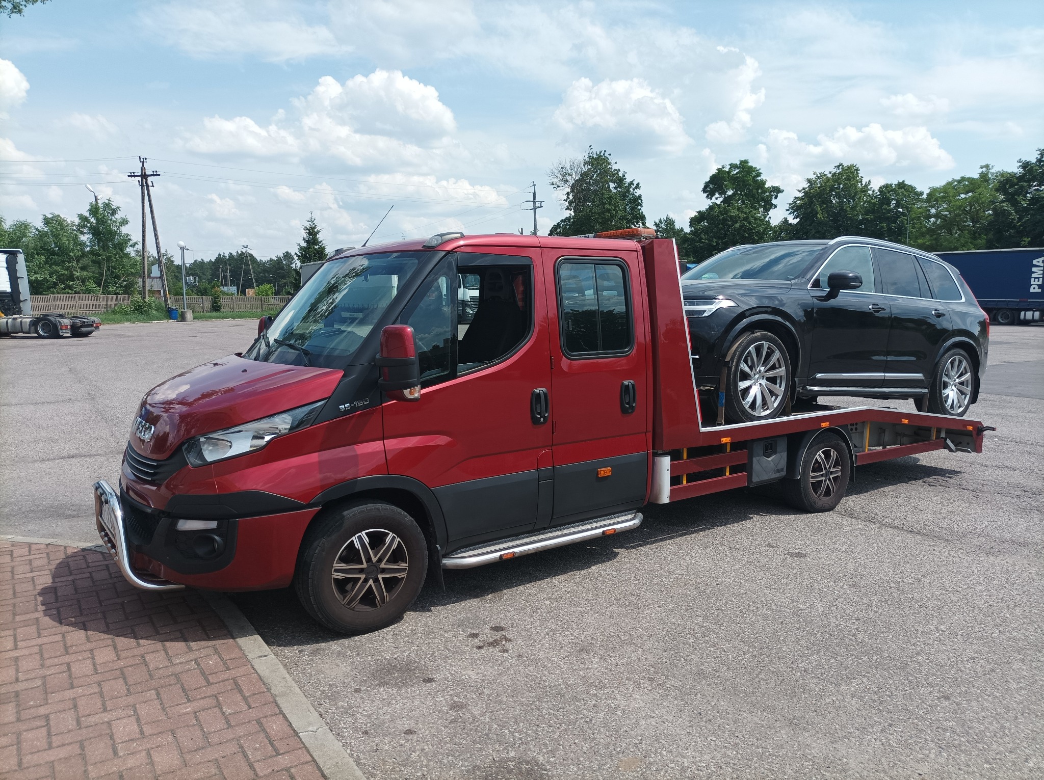 Czerwona ciężarówka pomocy drogowej Iveco z czarnym SUV-em Volvo na platformie, zaparkowana na parkingu z kostki brukowej w słoneczny dzień.