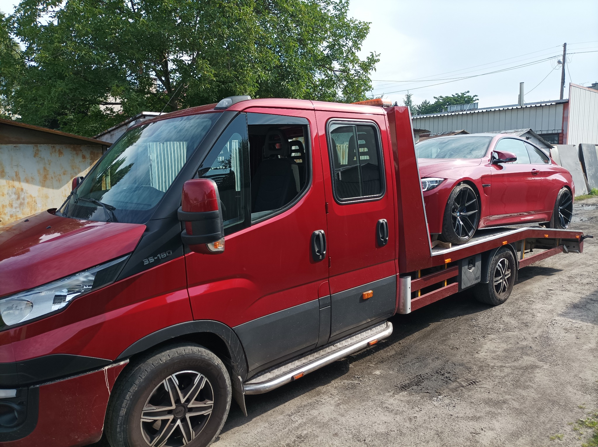 Czerwona platforma transportowa Iveco 35-180 przewozi czerwone sportowe coupe na tle budynku gospodarczego i zieleni drzew.