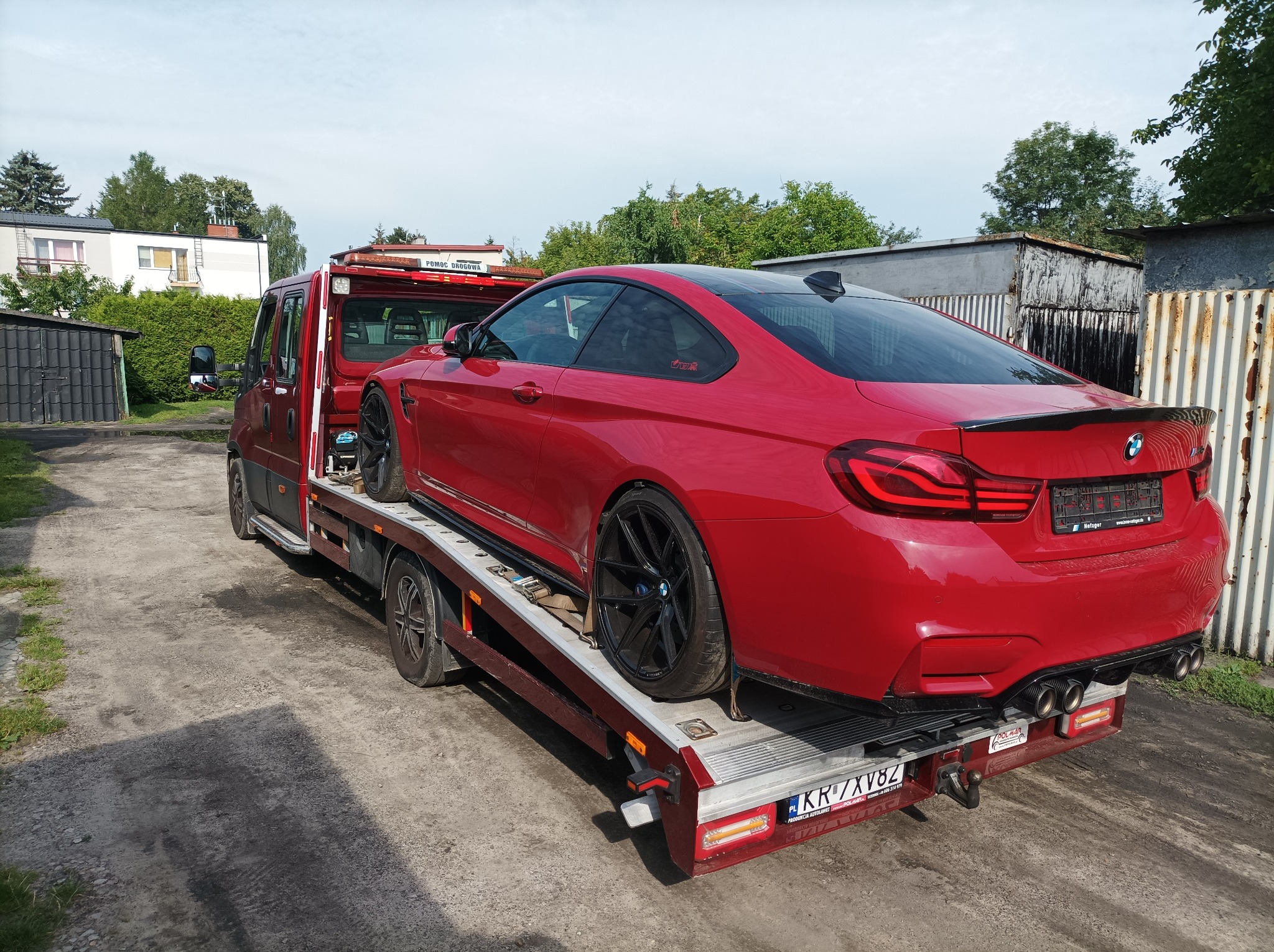 Czerwone sportowe BMW M4 na platformie pojazdu pomocy drogowej, widok z tyłu, na tle budynku i drzew.