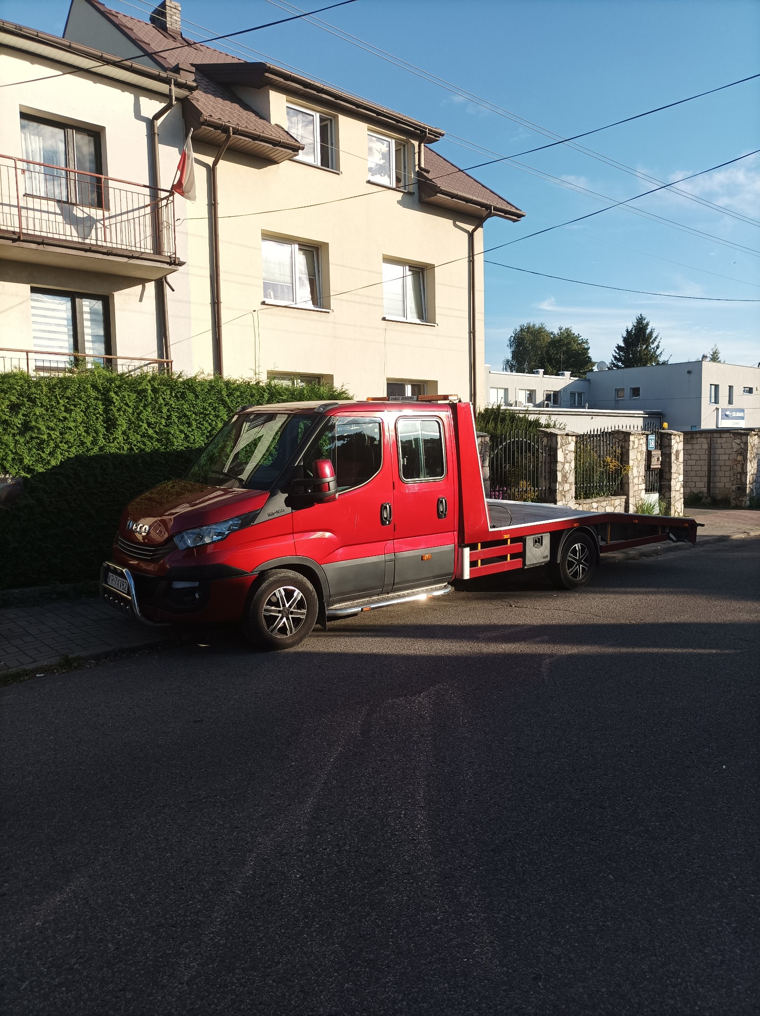 Czerwony samochód do transportu pojazdów marki Iveco zaparkowany na ulicy, z widocznym budynkiem mieszkalnym w tle.