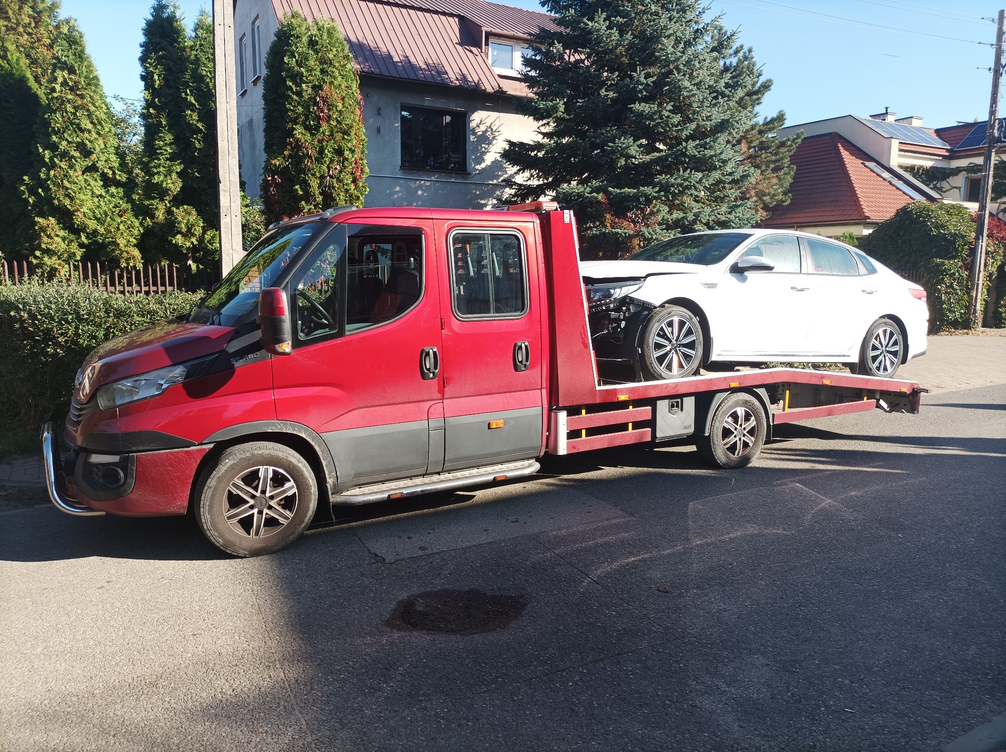 Czerwony pojazd transportowy z uszkodzonym białym samochodem osobowym na platformie, zaparkowany na ulicy w słoneczny dzień, z widocznymi domami i zielenią w tle.