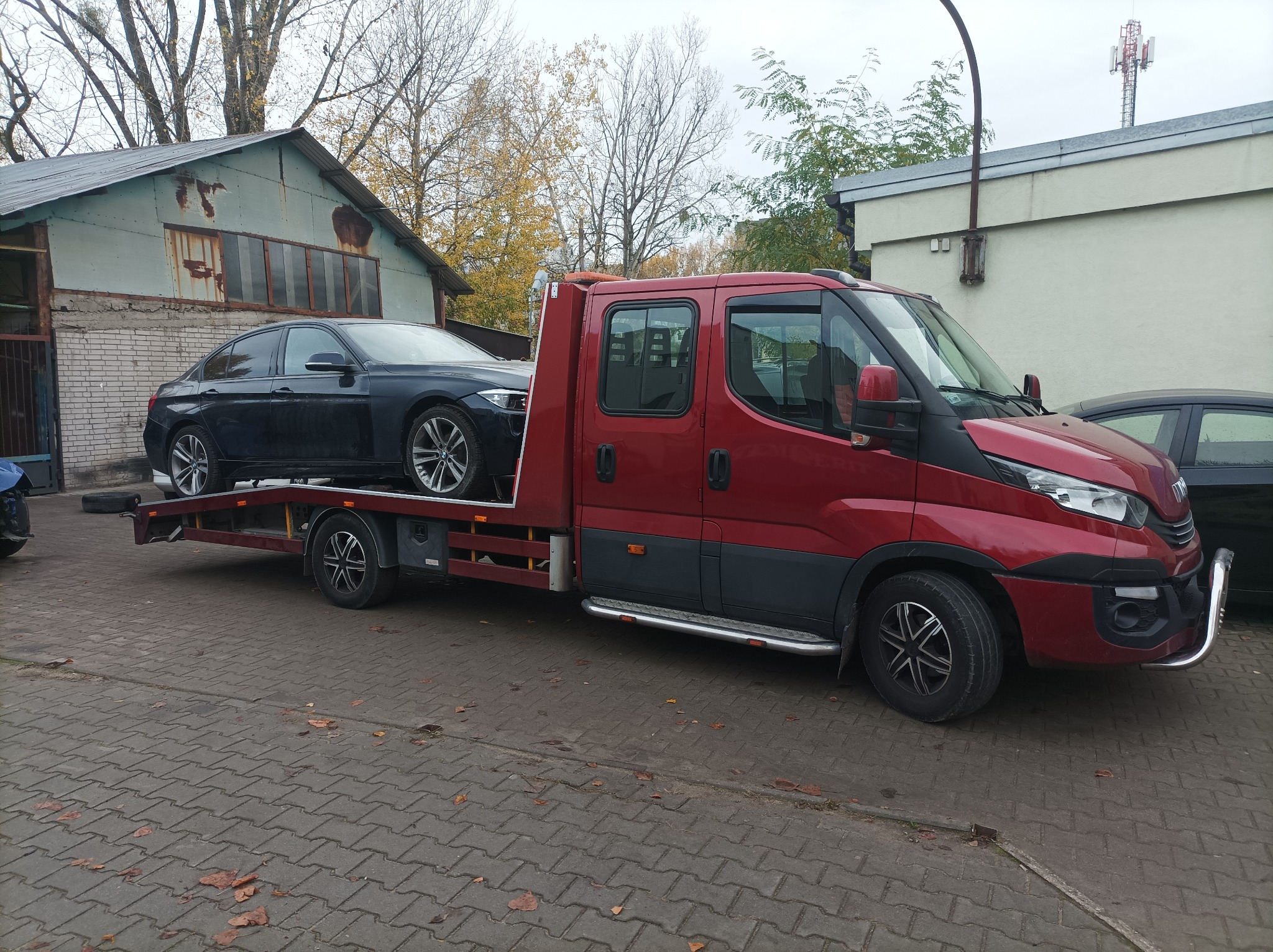 Czerwona laweta Iveco z załadowanym czarnym samochodem osobowym na tle zabudowań w Warszawie. Widoczne jesienne liście na chodniku.