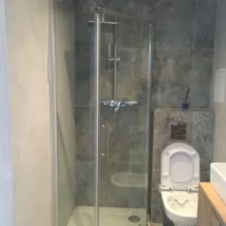 Nowoczesna łazienka z kabiną prysznicową ze szkła, toaletą podwieszaną i umywalką na drewnianej szafce, wykończona płytkami w odcieniach szarości i beżu.