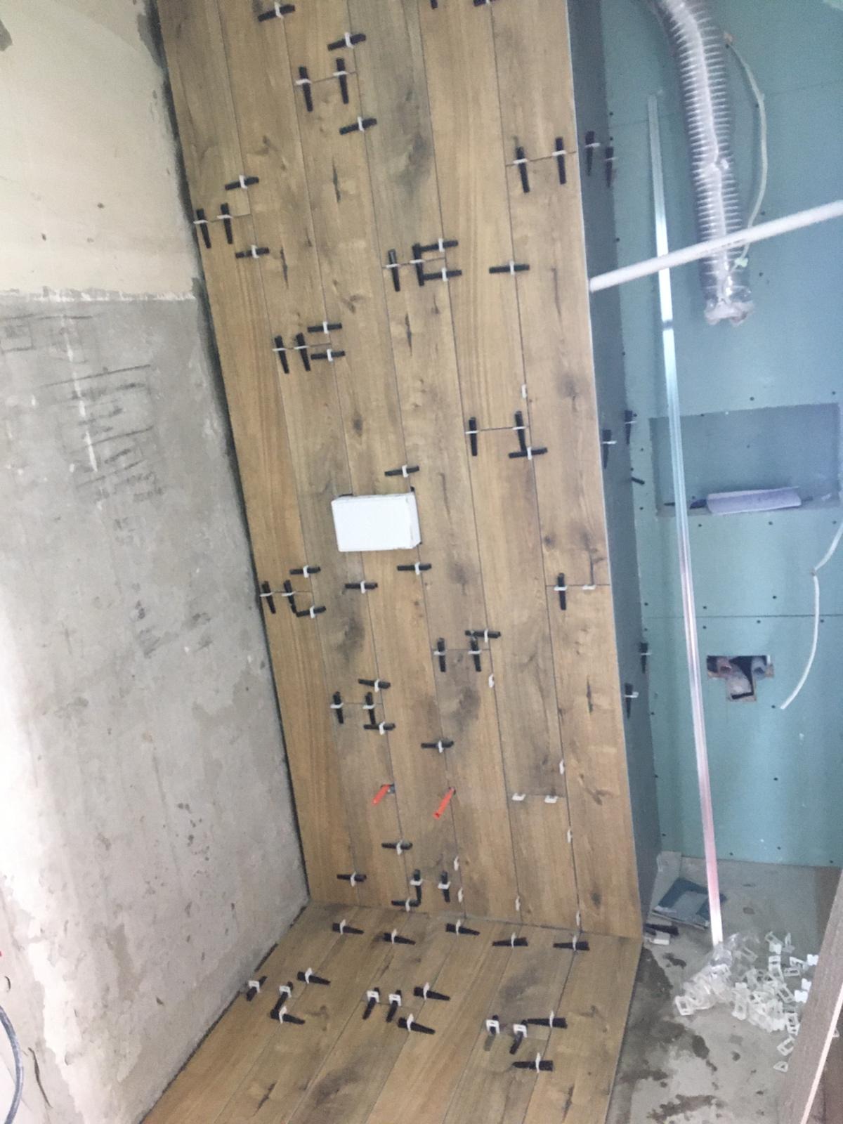 Montaż płytek imitujących drewno na ścianie w łazience, widoczne krzyżyki dystansowe i poziomica, surowa ściana obok.