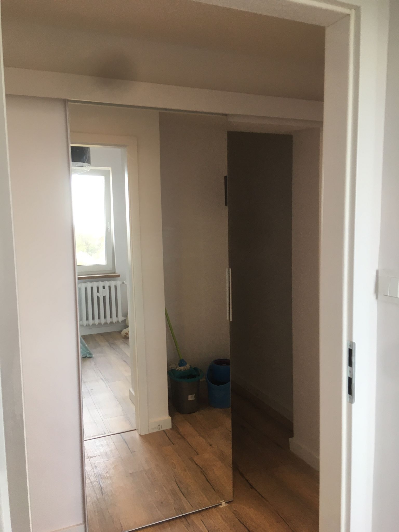 Zamontowane przesuwne drzwi z lustrem, widok na odnowione wnętrze z drewnianą podłogą i grzejnikiem pod oknem, częściowo widoczne akcesoria do sprzątania.