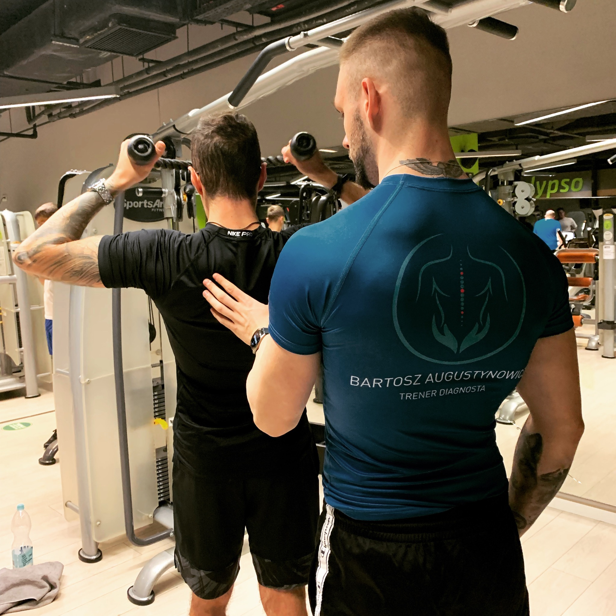 Trener personalny z tatuażami na ramieniu, w niebieskiej koszulce z logo i napisem 'Bartosz Augustynowicz Trener Diagnosta', pomaga mężczyźnie w czarnej koszulce ćwiczyć na maszynie do ćwiczeń...