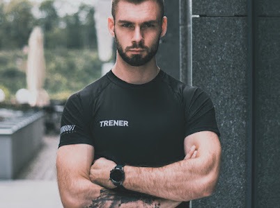 Mężczyzna z brodą w czarnej koszulce z napisem 'TRENER', stojący na zewnątrz z założonymi rękami, widoczny zegarek na nadgarstku i tatuaż na przedramieniu.