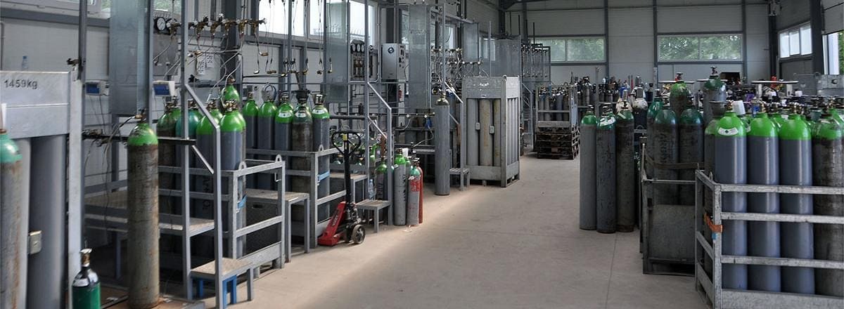 Magazyn butli z gazami technicznymi, głównie szare butle z zielonymi górnymi częściami, ustawione w metalowych regałach i kontenerach, z widoczną wagą 1459kg na jednym z regałów.