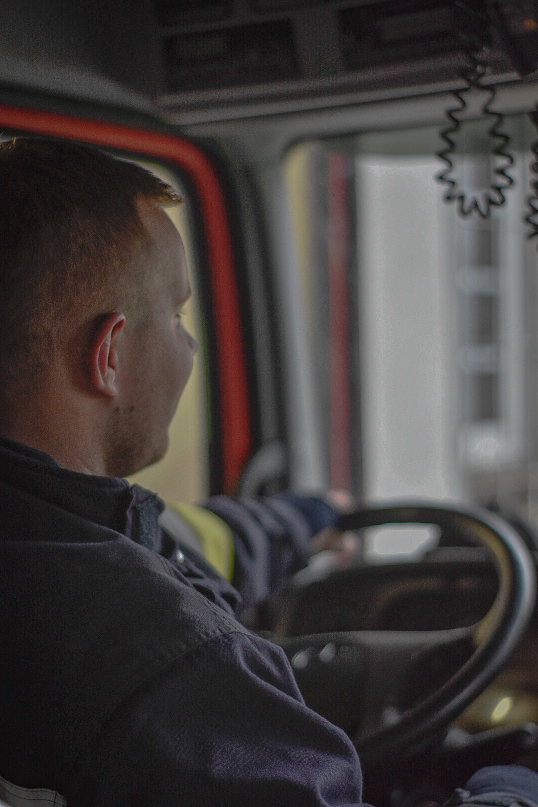 Mężczyzna w mundurze siedzi za kierownicą pojazdu, prawdopodobnie strażackiego, widok zza ramienia na profil twarzy i kierownicę.