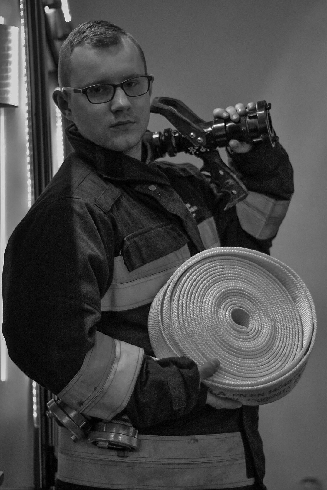 Strażak w okularach, w pełnym umundurowaniu, trzyma zwinięty wąż strażacki i dyszę, zdjęcie czarno-białe.