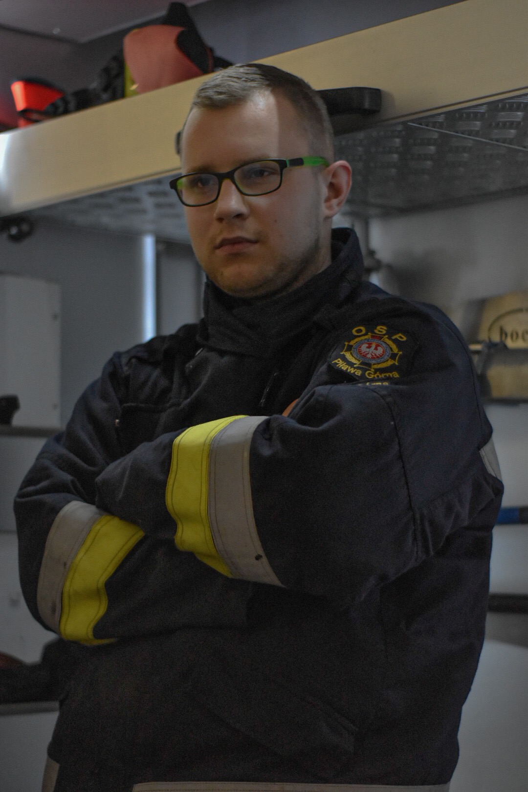 Portret strażaka w okularach, w mundurze z naszywką OSP Piława Górna, stojącego na tle wnętrza wozu strażackiego.