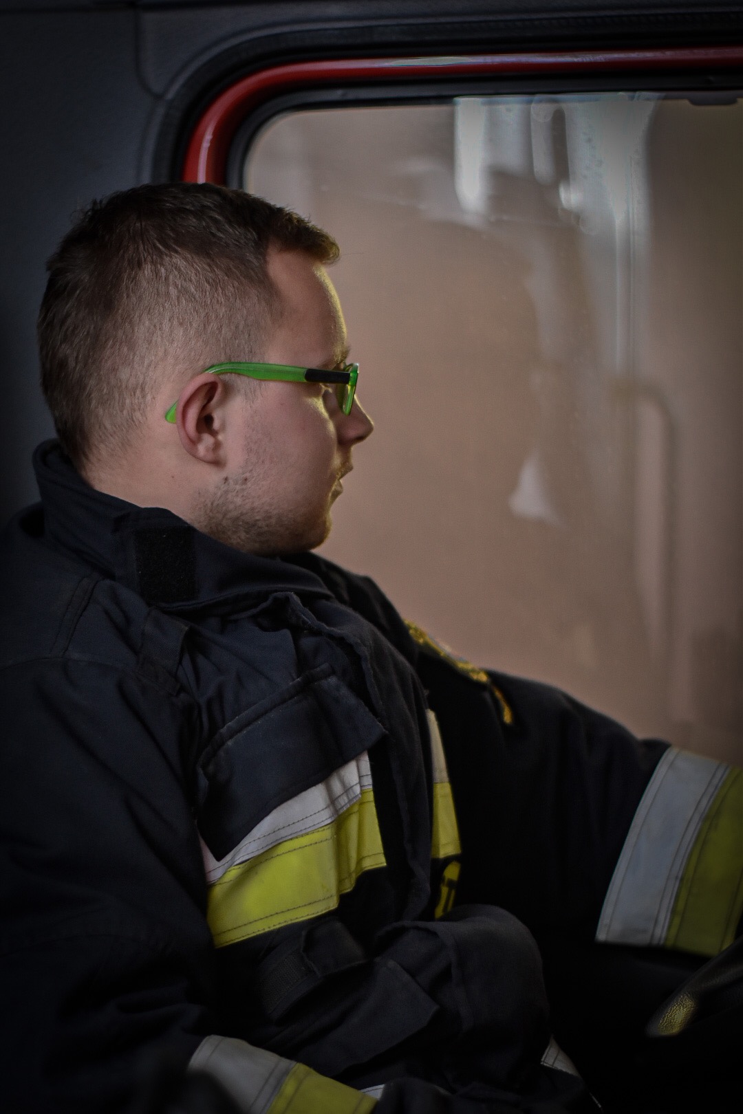 Strażak w mundurze, z zielonymi okularami, siedzi w kabinie wozu strażackiego, spoglądając przez okno.