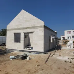 Wznoszenie ścian parterowego domu z białych bloczków, widoczne otwory okienne i drzwiowe, teren budowy z materiałami i narzędziami, w tle zabudowa jednorodzinna i drzewa iglaste.