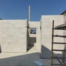 Wnętrze budynku w trakcie budowy, ściany wymurowane z białych bloczków, widoczne otwory okienne, fragment rusztowania i drewniany słupek. Na podłodze leży bloczek.