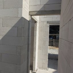 BUD-REX Michał Banasiak - Wnętrze wznoszonego domu z białych bloczków, widoczne fragmenty ścian z otworami drzwiowymi i nadprożami, świeży beton na podłodze, błękitne niebo w tle.