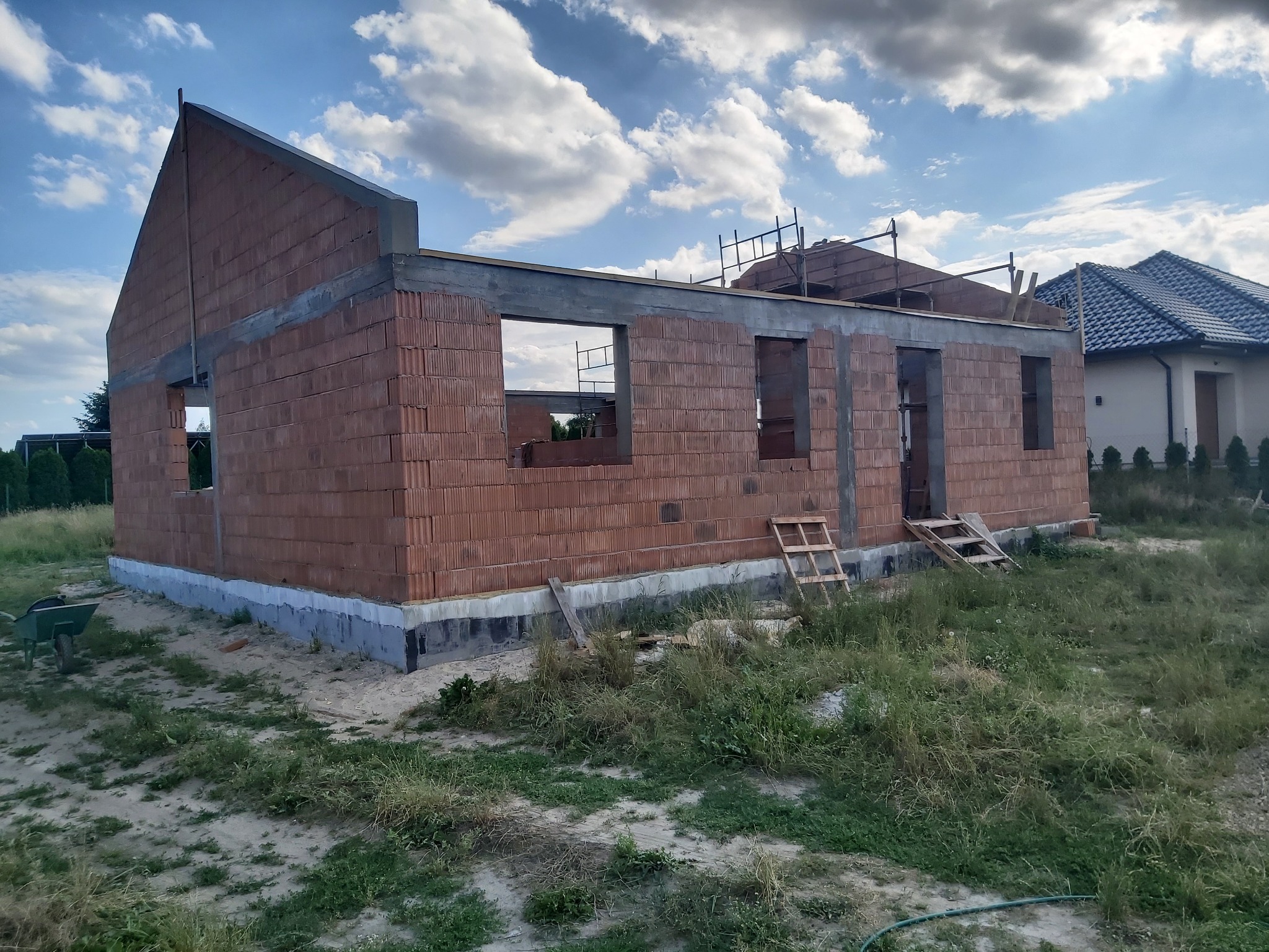 Wznoszenie ścian parterowego domu z czerwonej cegły ceramicznej Porotherm, widoczne otwory okienne i drzwiowe, szare nadproża, częściowo wykonane fundamenty, w tle błękitne niebo z białymi chmurami.
