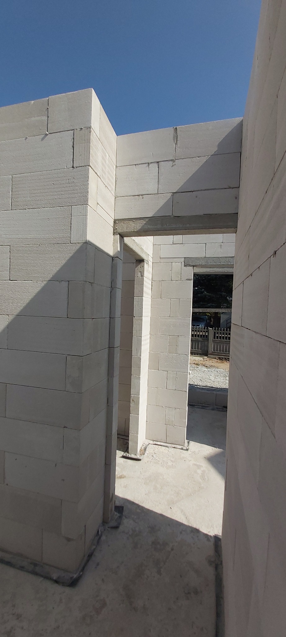 Wnętrze wznoszonego domu z białych bloczków, widoczne fragmenty ścian z otworami drzwiowymi i nadprożami, świeży beton na podłodze, błękitne niebo w tle.