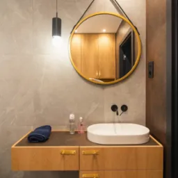 Nowoczesna łazienka z okrągłym lustrem w złotej ramie, wiszącą szafką z umywalką i ciemnymi akcentami, takimi jak bateria i lampa. Szare płytki imitujące marmur na ścianach.