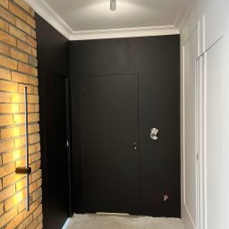 Daniel Glapa D.M.G. House Finish - Nowoczesne wykończenie wnętrza: czarne ściany z oświetleniem LED przy ceglanej ścianie, białe listwy przysufitowe, minimalistyczne drzwi i surowe wykończenie podłogi.