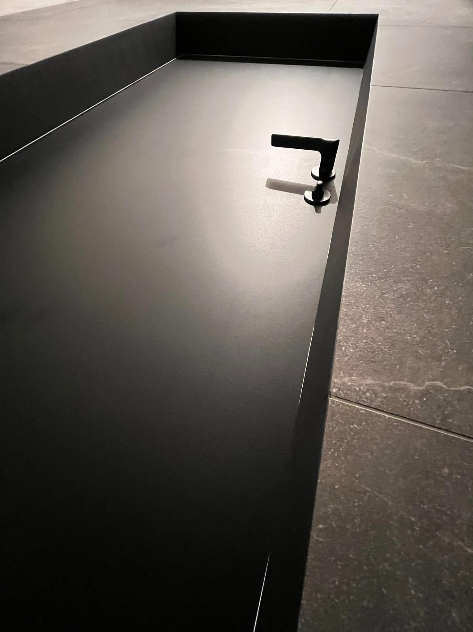 Czarny, minimalistyczny blat umywalkowy z ciemną armaturą i szarą płytką podłogową. Nowoczesne wykończenie łazienki, detal.