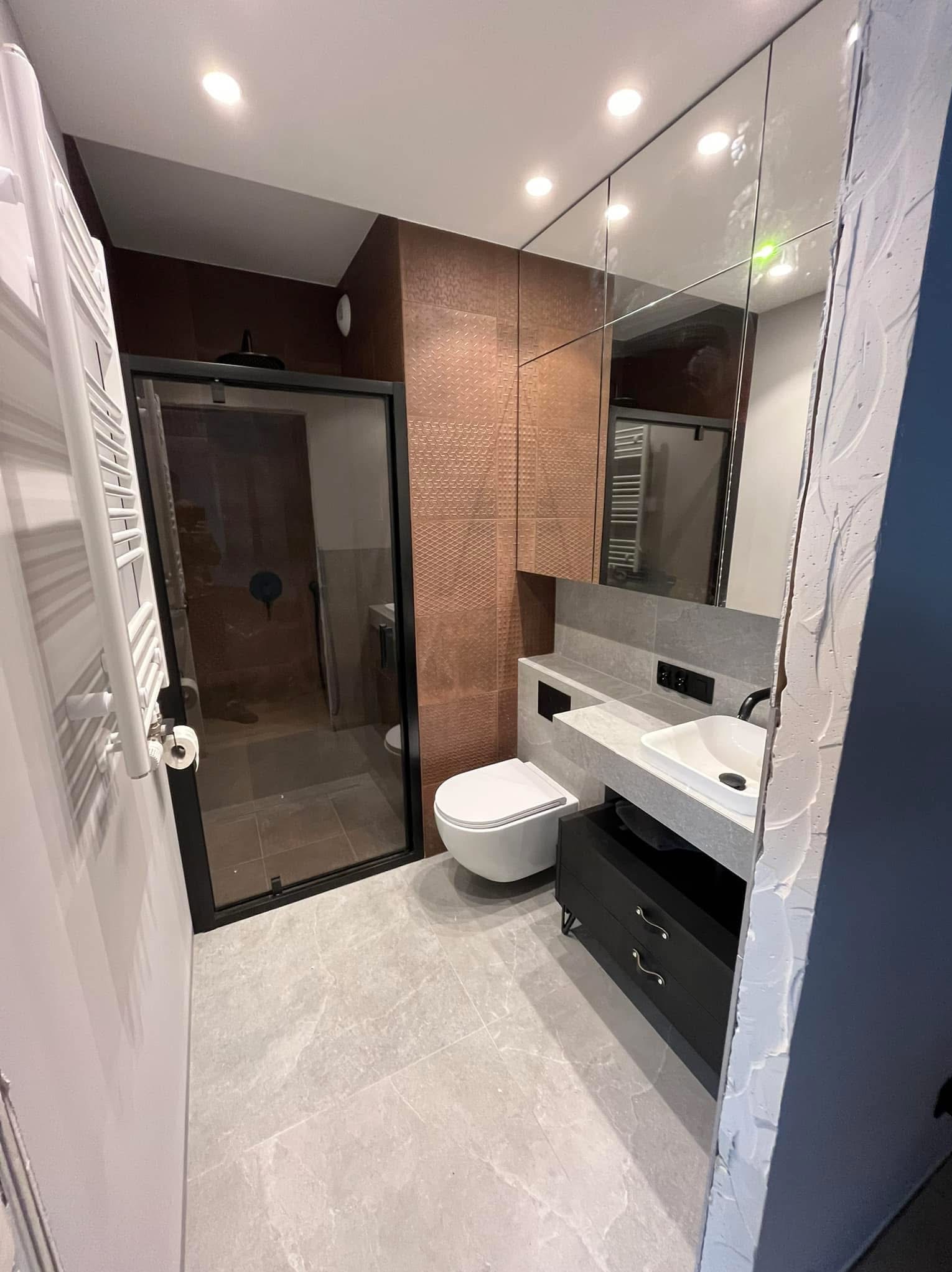 Nowoczesna łazienka po remoncie: kabina prysznicowa z czarną ramą, toaleta, umywalka z szafką i duże lustro. Brązowe płytki na ścianie, szare płytki na podłodze.