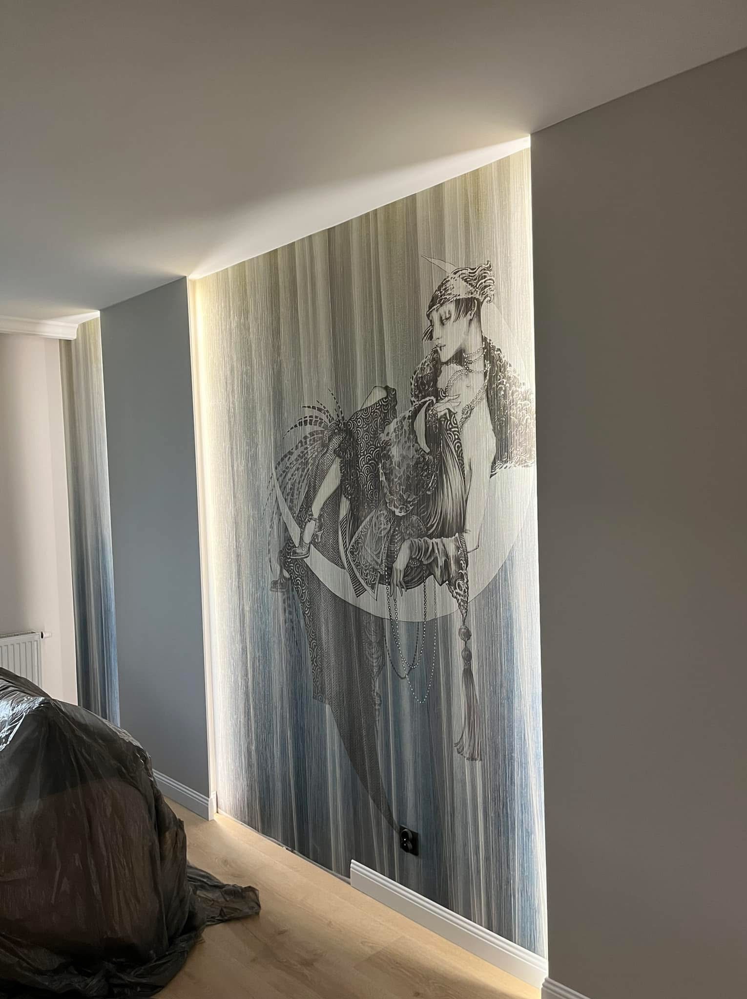 Ściana z tapetą w paski zdobiona czarno-białym rysunkiem kobiety, oświetlona pionowo z góry. Elementy wykończenia podłogi i mebel pod folią ochronną.