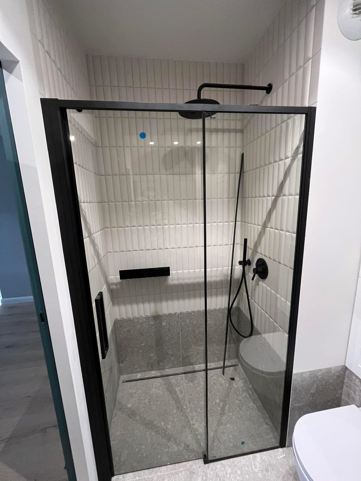 Nowoczesna kabina prysznicowa z czarnymi ramami i armaturą. Białe, pionowe płytki na ścianach, szare płytki na podłodze. Minimalistyczny design.