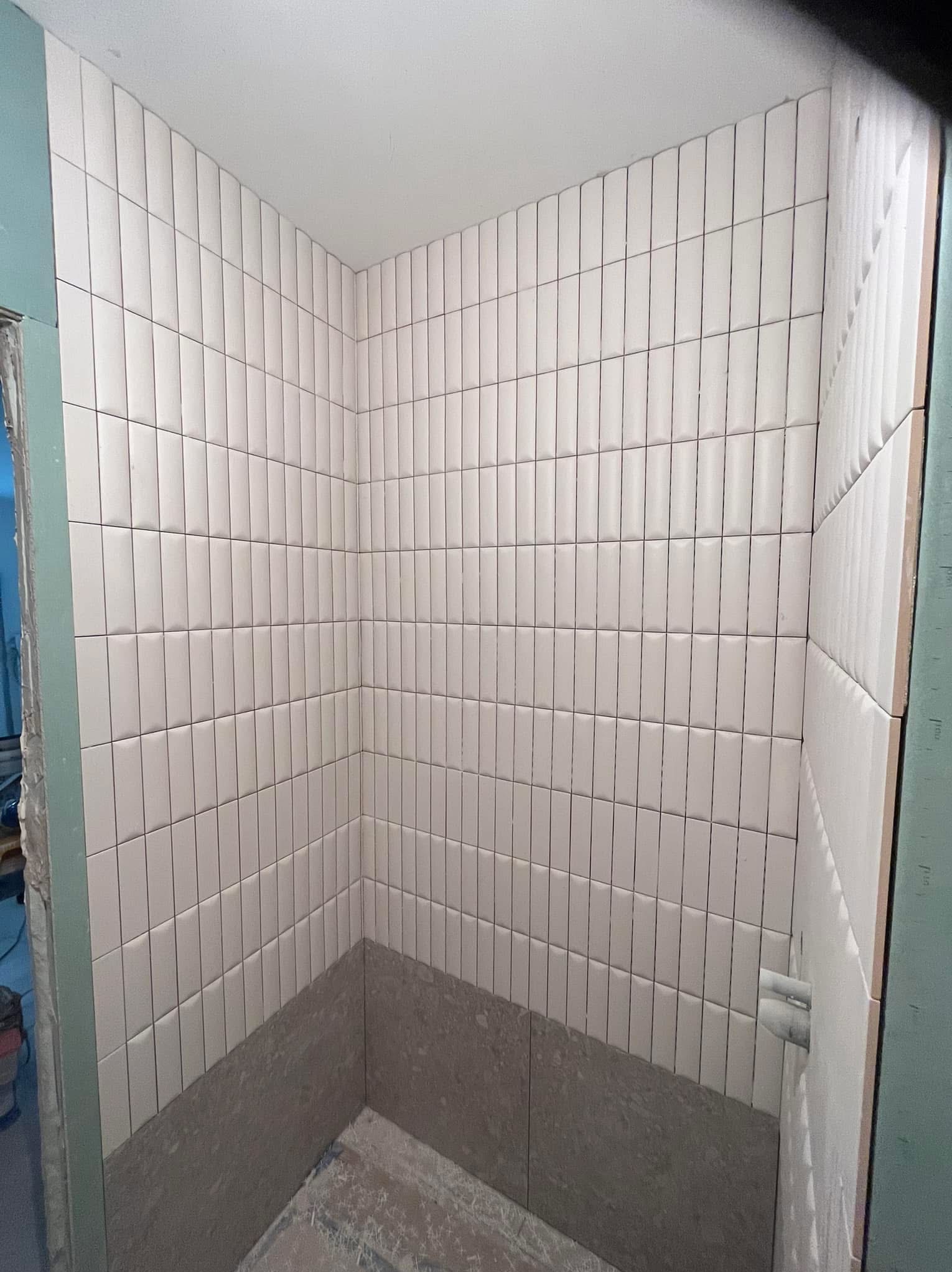 Ściana w trakcie wykańczania z ułożonymi białymi, pionowymi płytkami w kształcie prostokątów. Dolna część ściany przygotowana pod dalsze prace wykończeniowe.