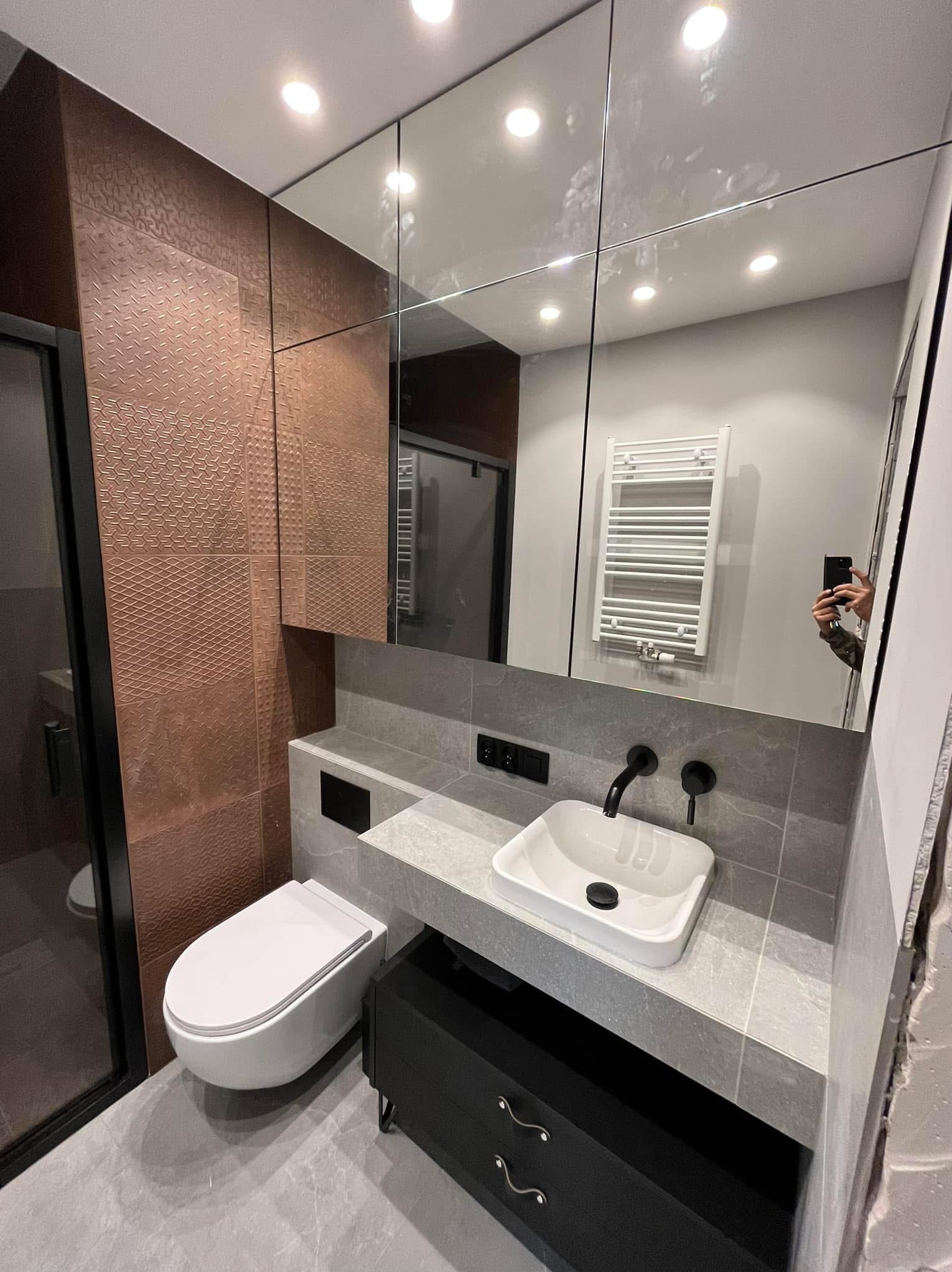 Nowoczesna łazienka z miedzianymi płytkami strukturalnymi, szarą armaturą i dużym lustrem. Widoczny fragment kabiny prysznicowej i toaleta podwieszana.