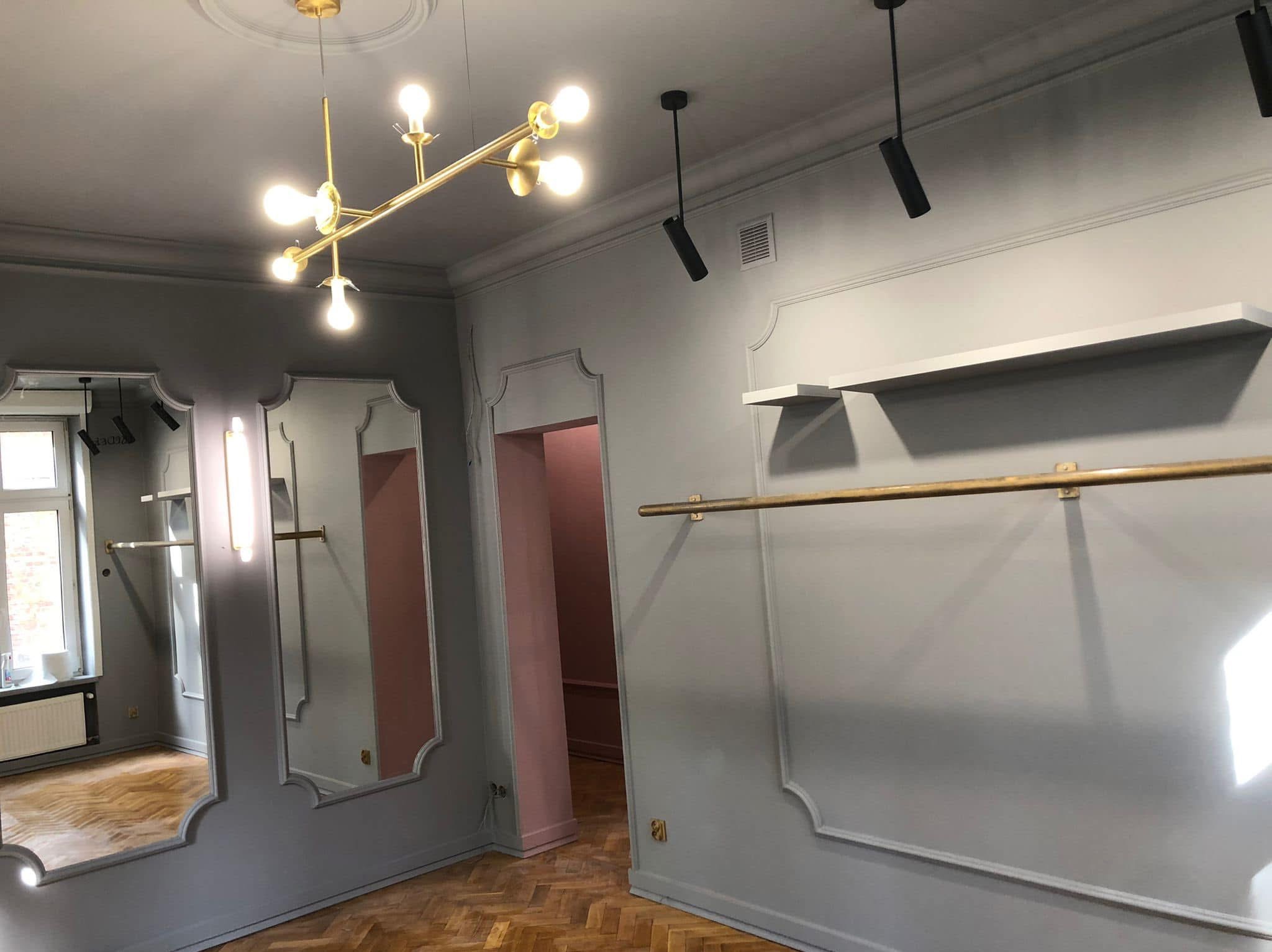 Pomalowane na szaro ściany z dekoracyjnymi listwami w sklepie odzieżowym, lustra w ozdobnych ramach, parkiet w jodełkę, złote lampy i karnisze.