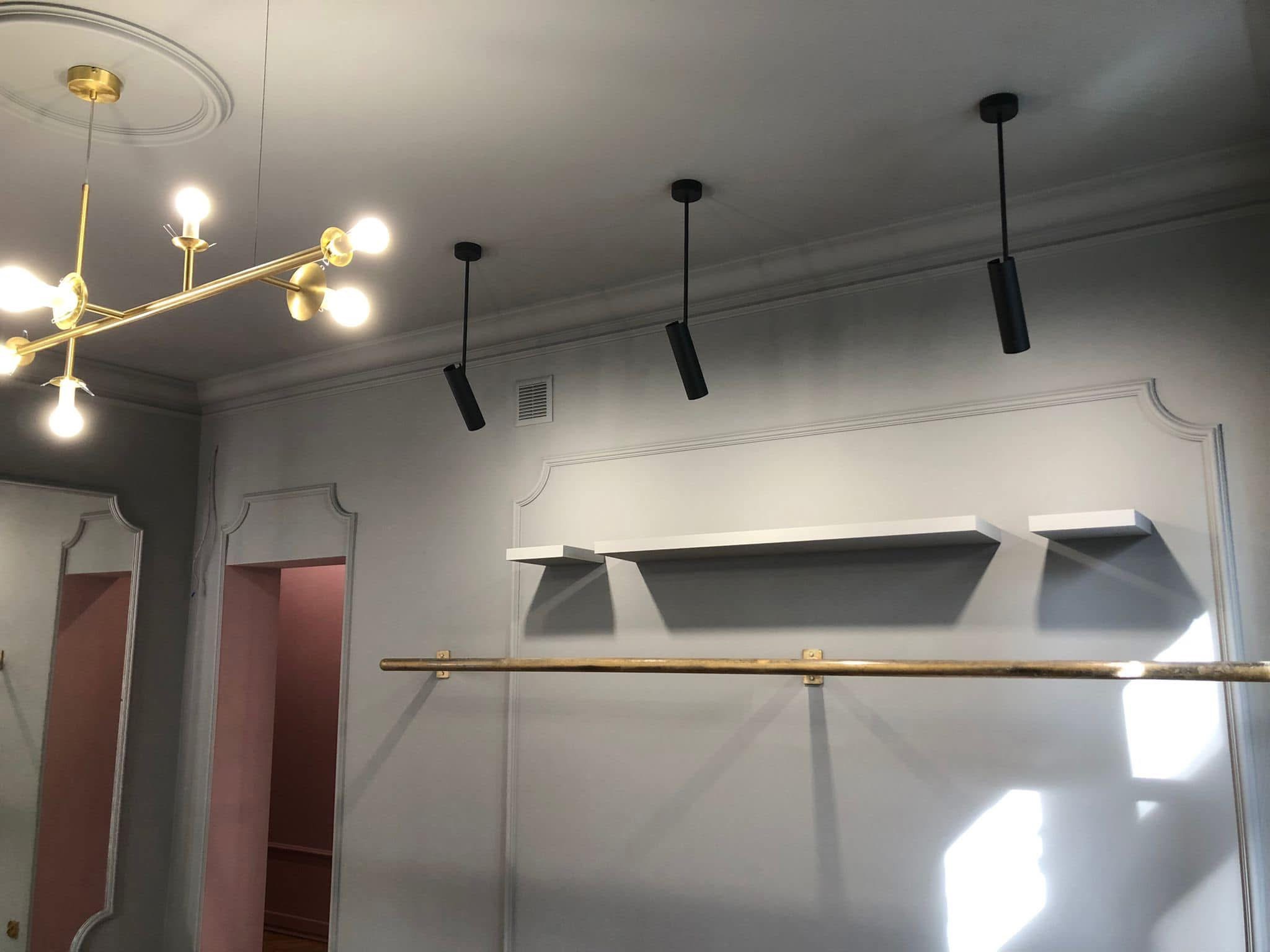Szare wnętrze butiku z dekoracyjną sztukaterią, złotym oświetleniem i metalowym drążkiem na ubrania. Widoczne półki i czarne reflektory punktowe.