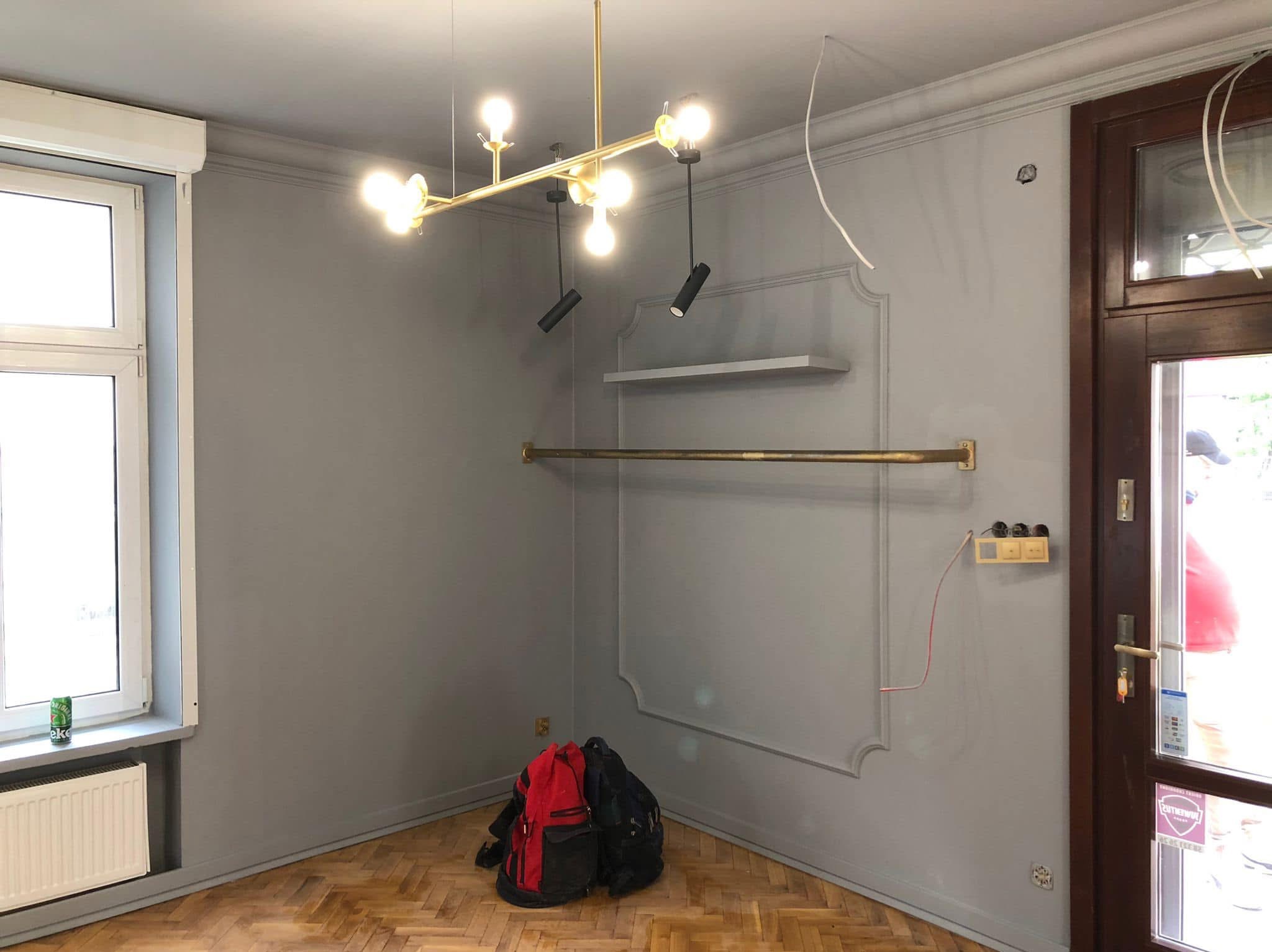Szare wnętrze pokoju z parkietem, złotą lampą i drążkiem na ubrania. Widoczne niedokończone instalacje elektryczne i torby na podłodze.