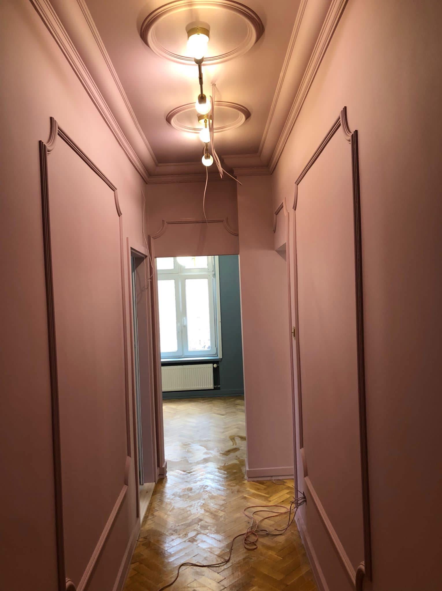 Korytarz po remoncie w odcieniach pudrowego różu. Ściany z dekoracyjnymi listwami, parkiet w jodełkę, sufit z rozetami i nowoczesnym oświetleniem. Widok na pokój z oknem.