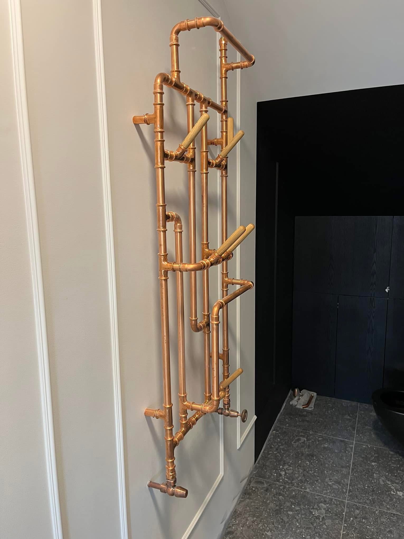 Miedziany grzejnik łazienkowy z drewnianymi wieszakami, zamontowany na szarej ścianie z dekoracyjnymi listwami. W tle czarne drzwi i fragment szarej podłogi.