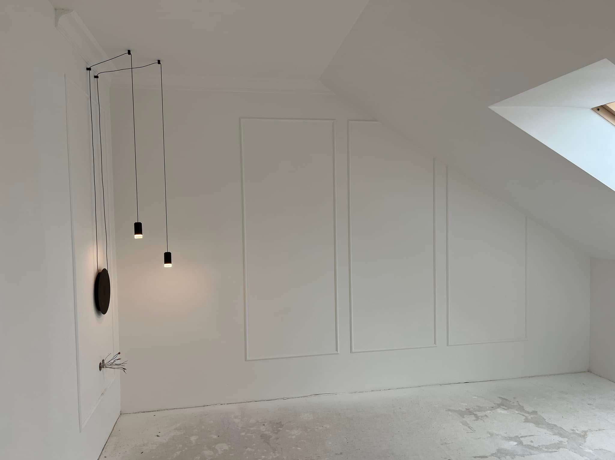 Białe ściany poddasza z dekoracyjnymi panelami i nowoczesnym oświetleniem. Widoczne kable elektryczne i surowe wykończenie podłogi.