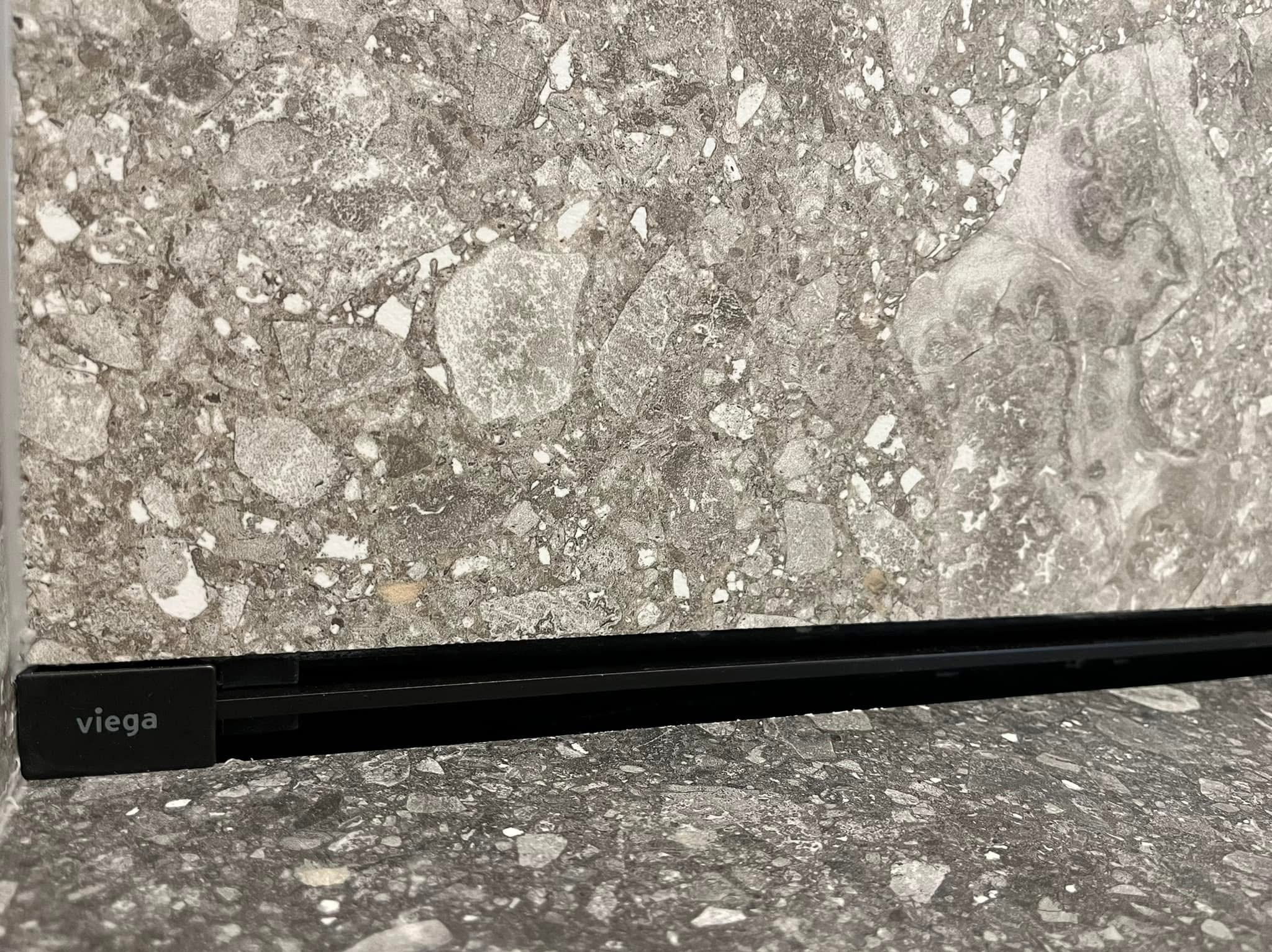 Szara, wzorzysta płytka z czarną listwą Viega. Detal wykończenia podłogi z bliska, ukazujący fakturę kamienia i precyzję montażu elementu wykończeniowego.