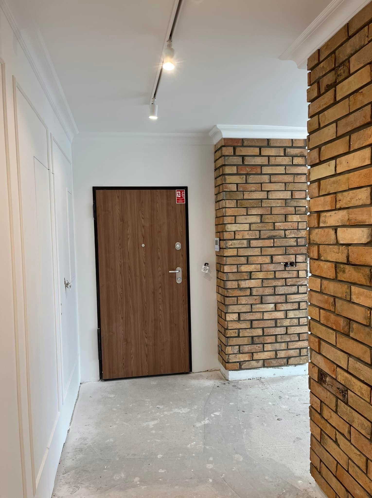 Korytarz po remoncie: ceglana kolumna i drzwi wejściowe. Białe ściany z dekoracyjnymi listwami, sufit z oświetleniem szynowym. Podłoga przygotowana do wykończenia.