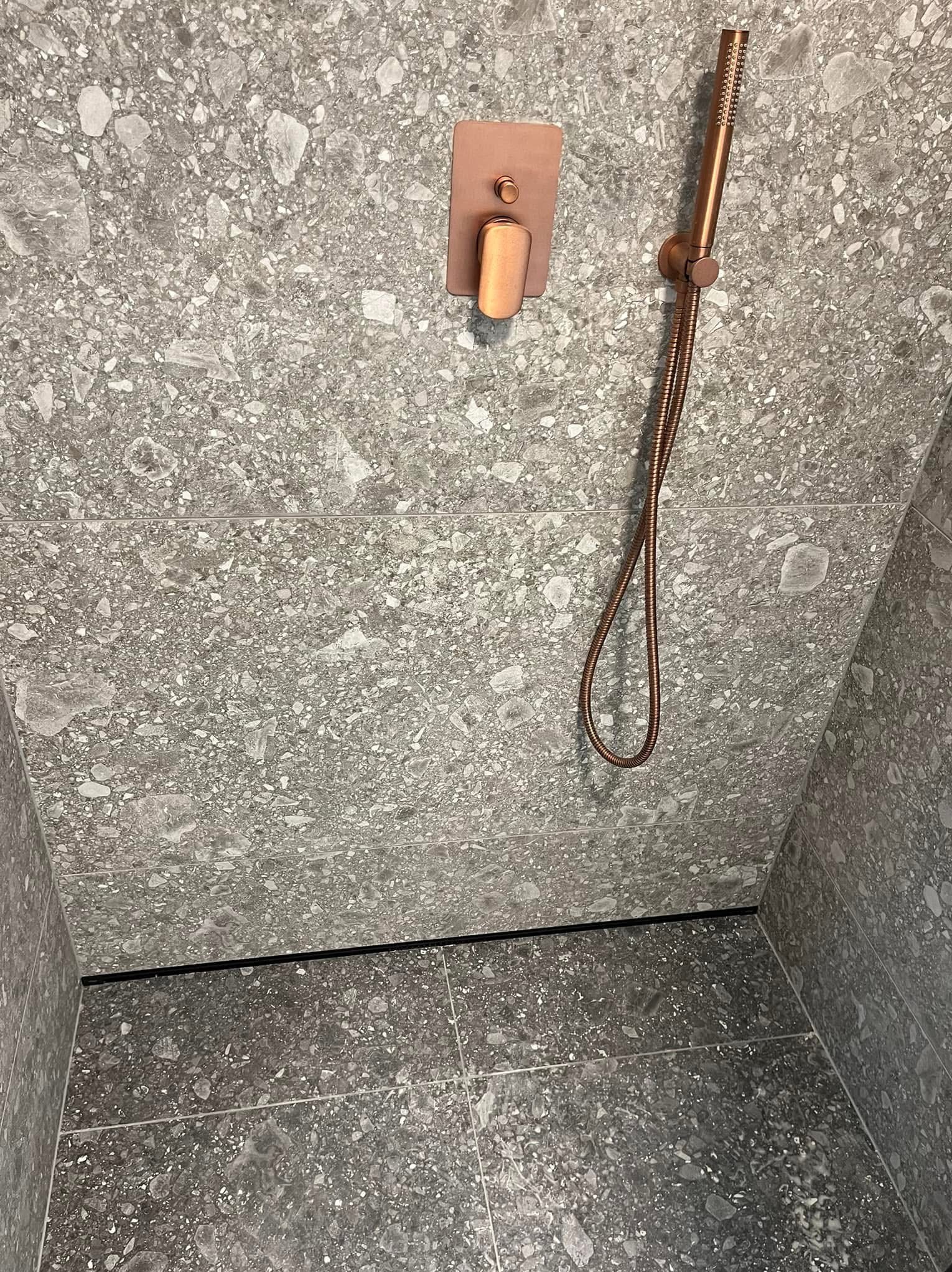 Wykończona kabina prysznicowa z szarymi płytkami lastryko i miedzianą armaturą. Minimalistyczny design, widok na fragment ściany i podłogi.